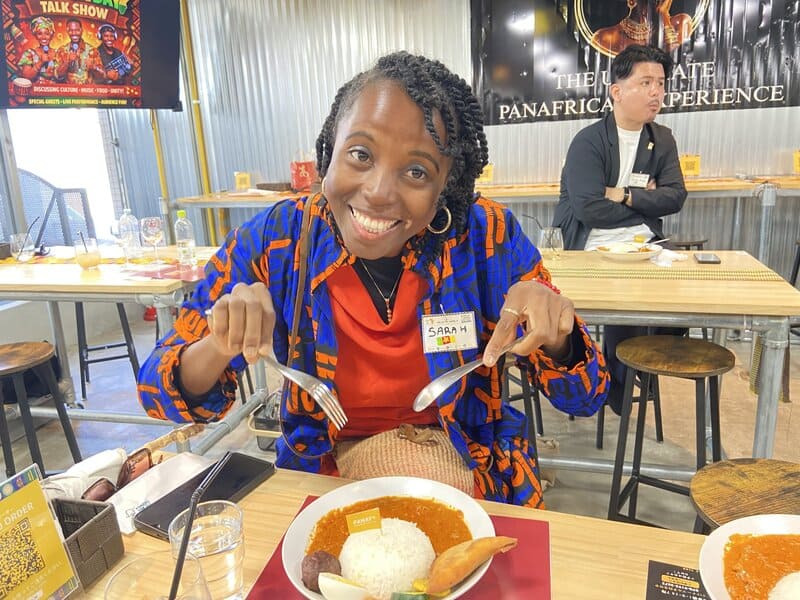 Sarah smiling while eating a traditional plate of rice and groundnut stew at PANAF Restaurant in Japan.