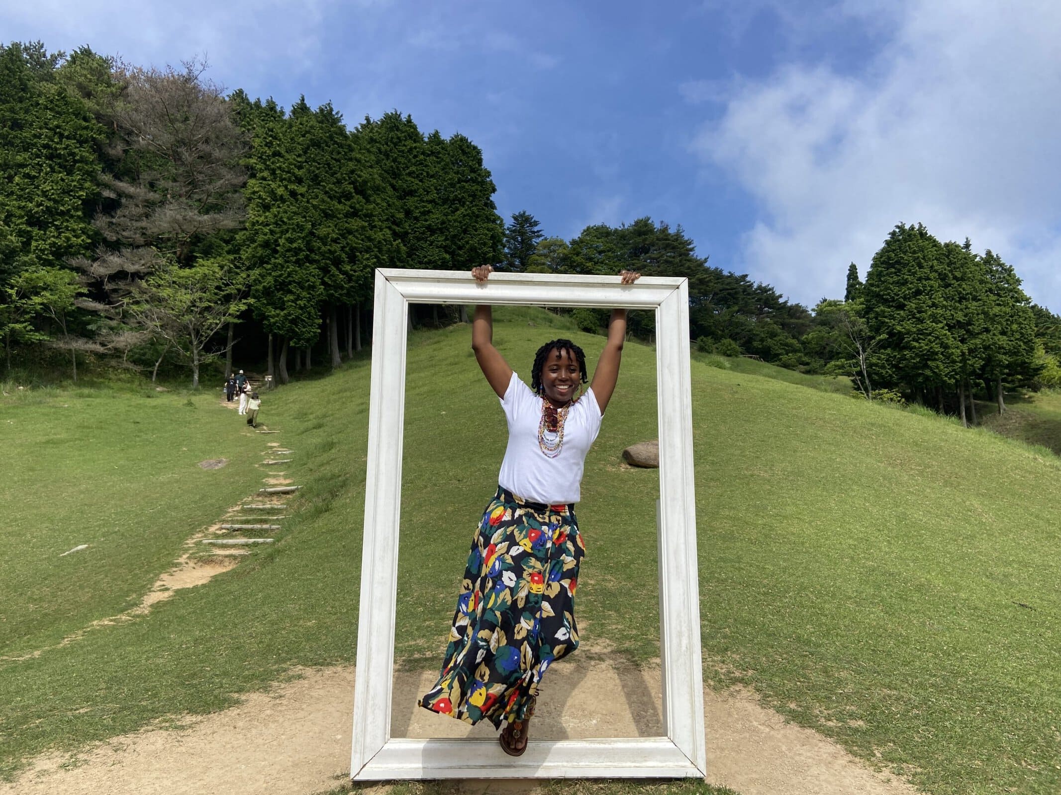 sarah namondo holding a large empty picture frame outdoors on a grassy hill.