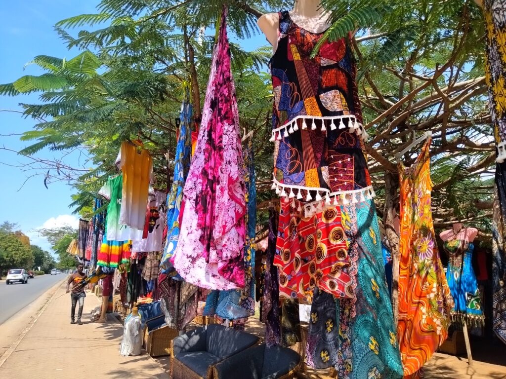 A vibrant display of colourful patterned Kenyan fabrics, dresses, and beachwear hanging from a wooden structure at a roadside market stall in Diani Beach
