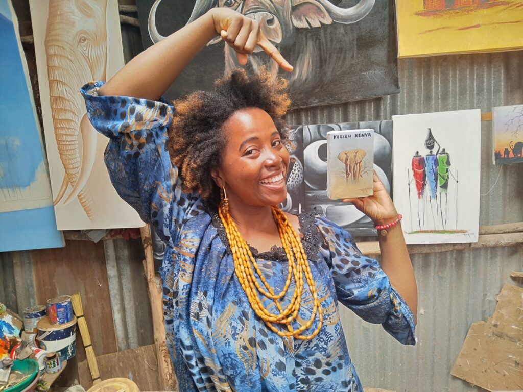 Sarah smiling and holding up a colourful, hand-painted portrait of herself while standing inside a local Diani Beach craft market.