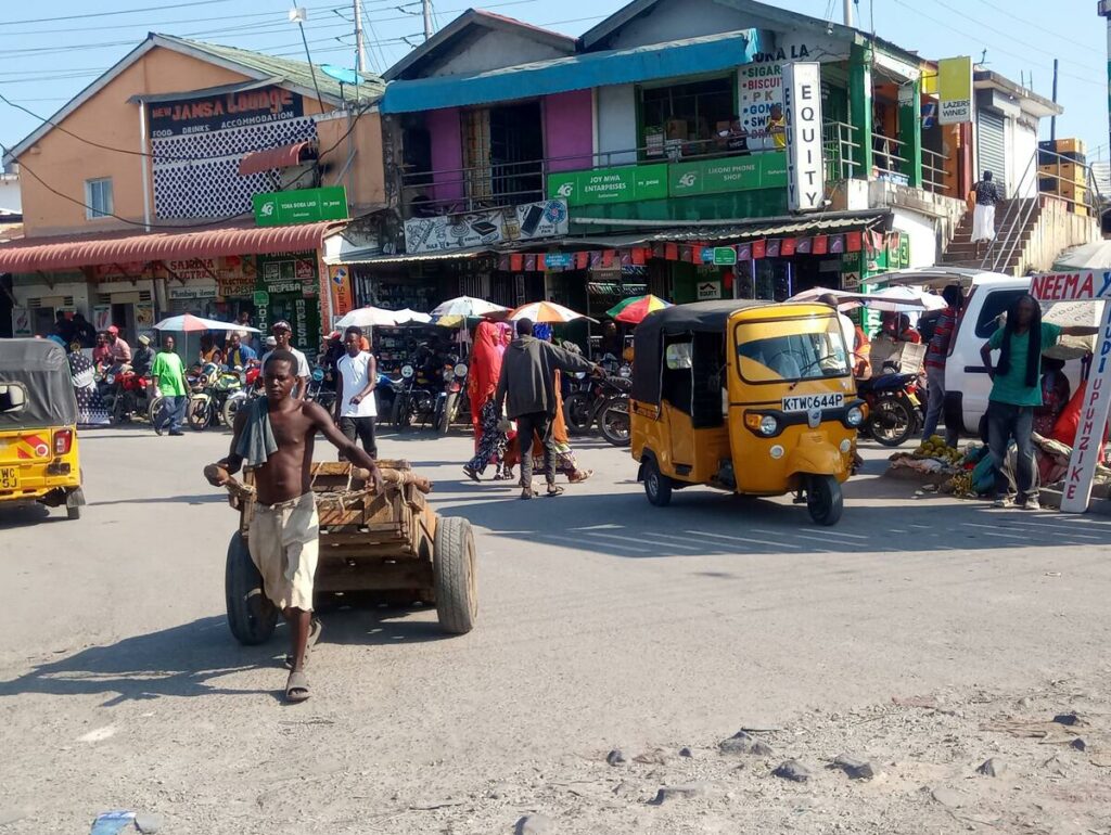 A busy street scene in Diani Beach, Kenya, featuring colourful tuk-tuks, local shops, and pedestrians moving along the road.