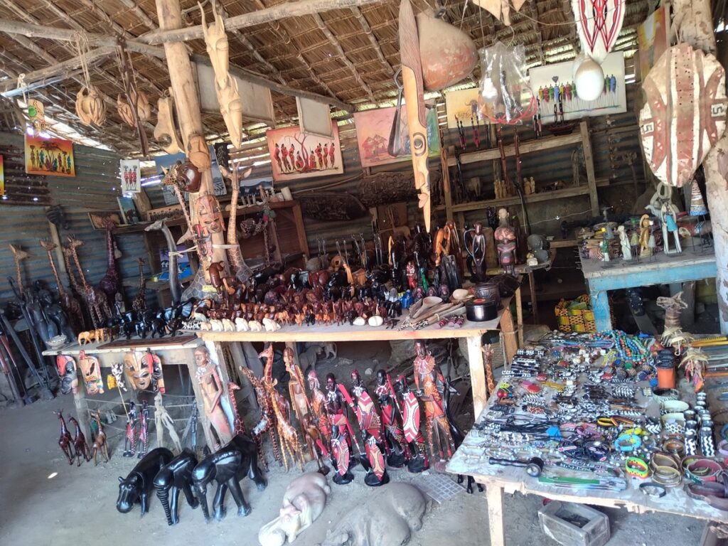 A collection of hand-carved wooden sculptures, including animal figures and decorative bowls, displayed for sale at an open-air Diani Beach craft stall.